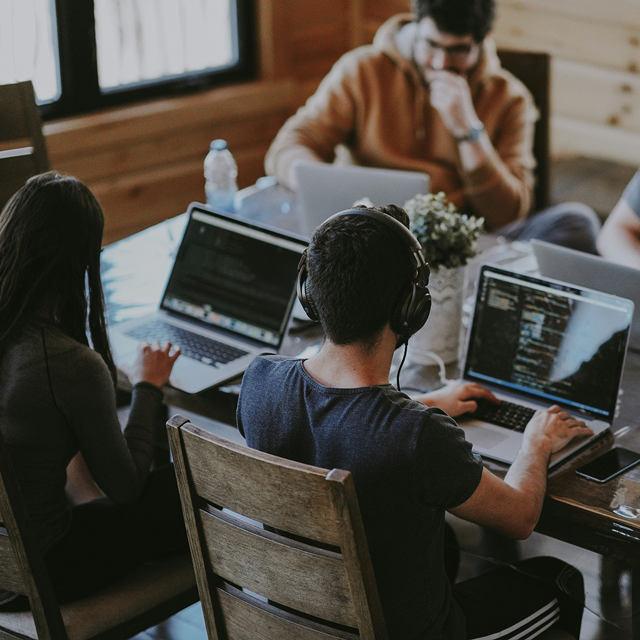 A photo of a group of young people sitting around a table while working on their laptops. 